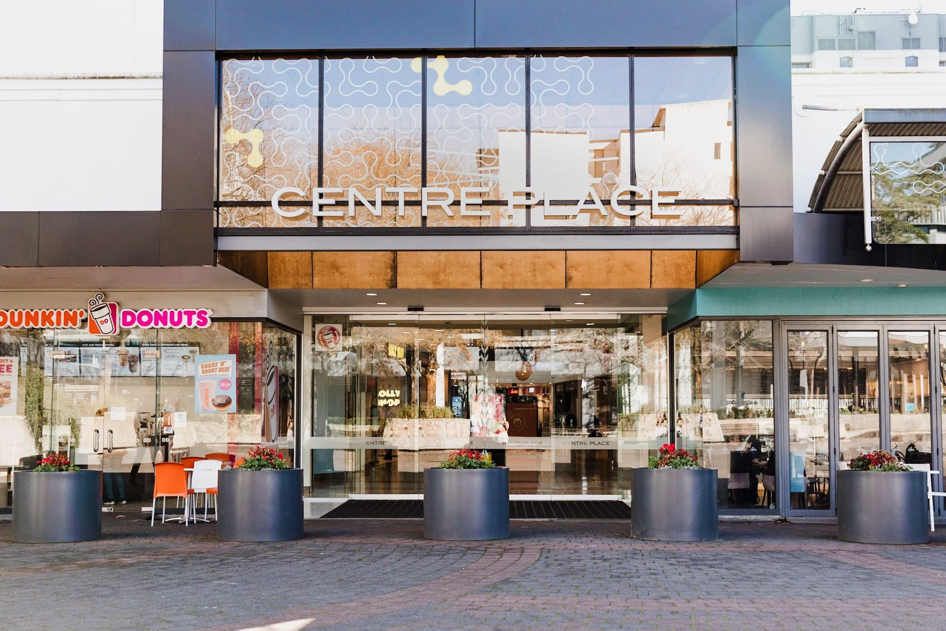 Shopping center entrance with glass storefronts and outdoor seating, including a Dunkin' Donuts café.