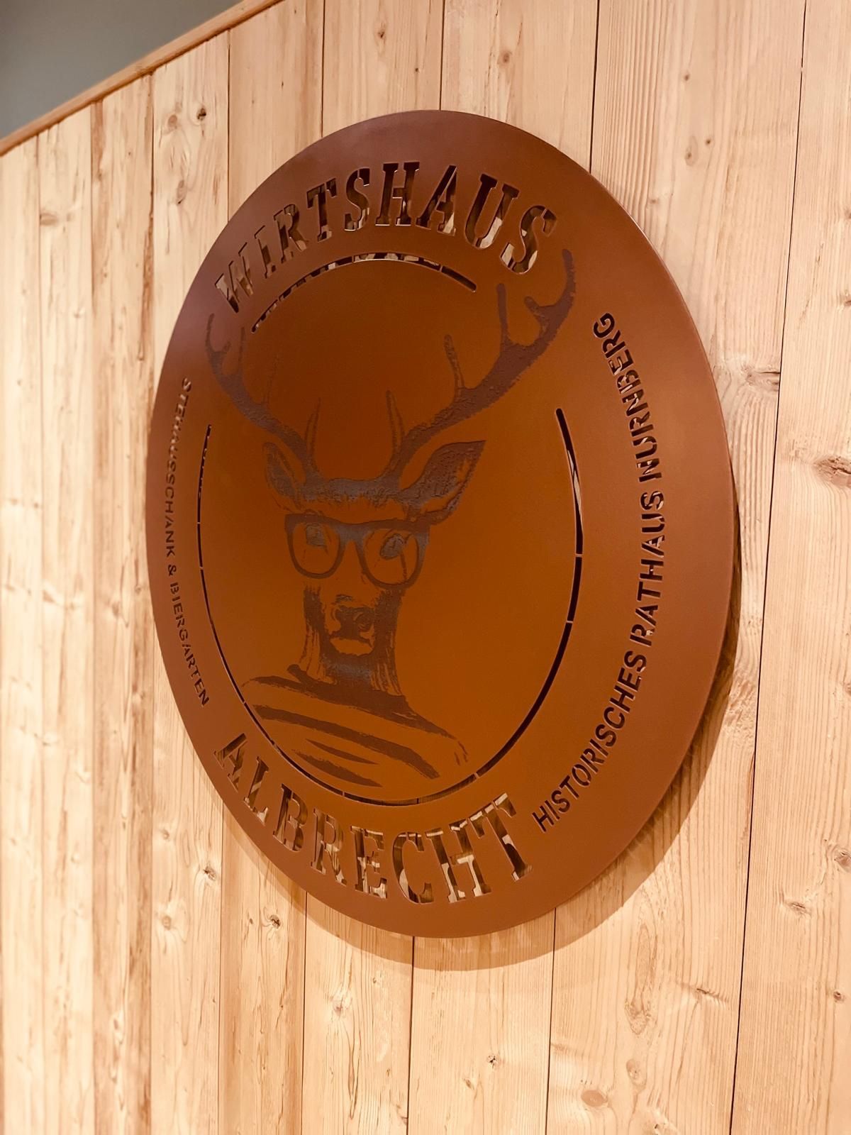 Logo in einer Metallplatte ausgefräst auf einer Wand aus Holz hängend.