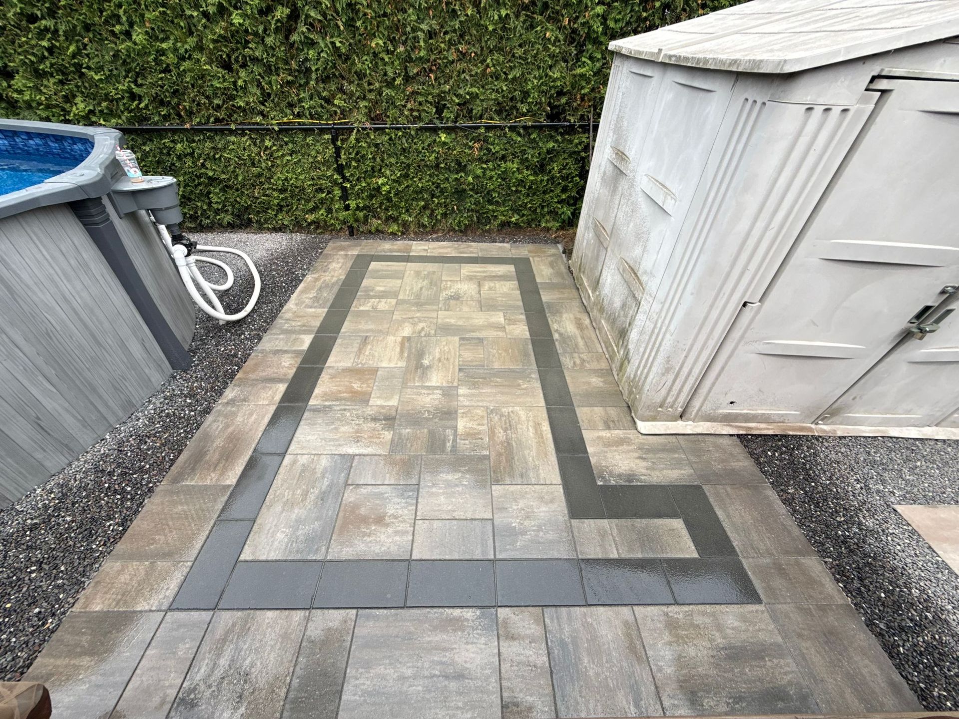 Patio avec pavés gris, bordure noire et cabanon à côté d'une piscine hors terre et gravier.