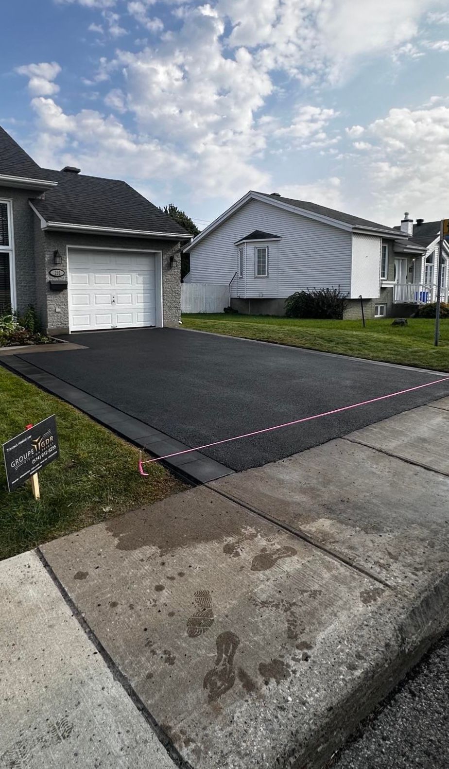 Allée en asphalte noir nouvellement pavée à côté d'une pelouse herbeuse et d'un trottoir en béton, avec des maisons en arrière-plan.