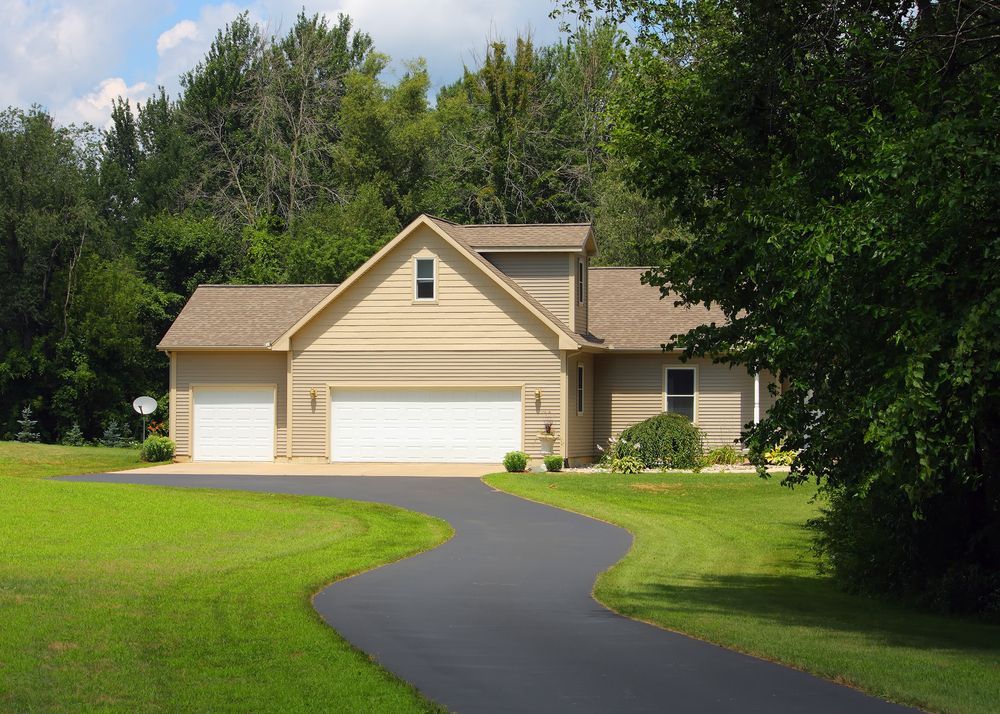 Maison beige avec garage pour trois voitures, allée noire incurvée, pelouse verte et arbres environnants.