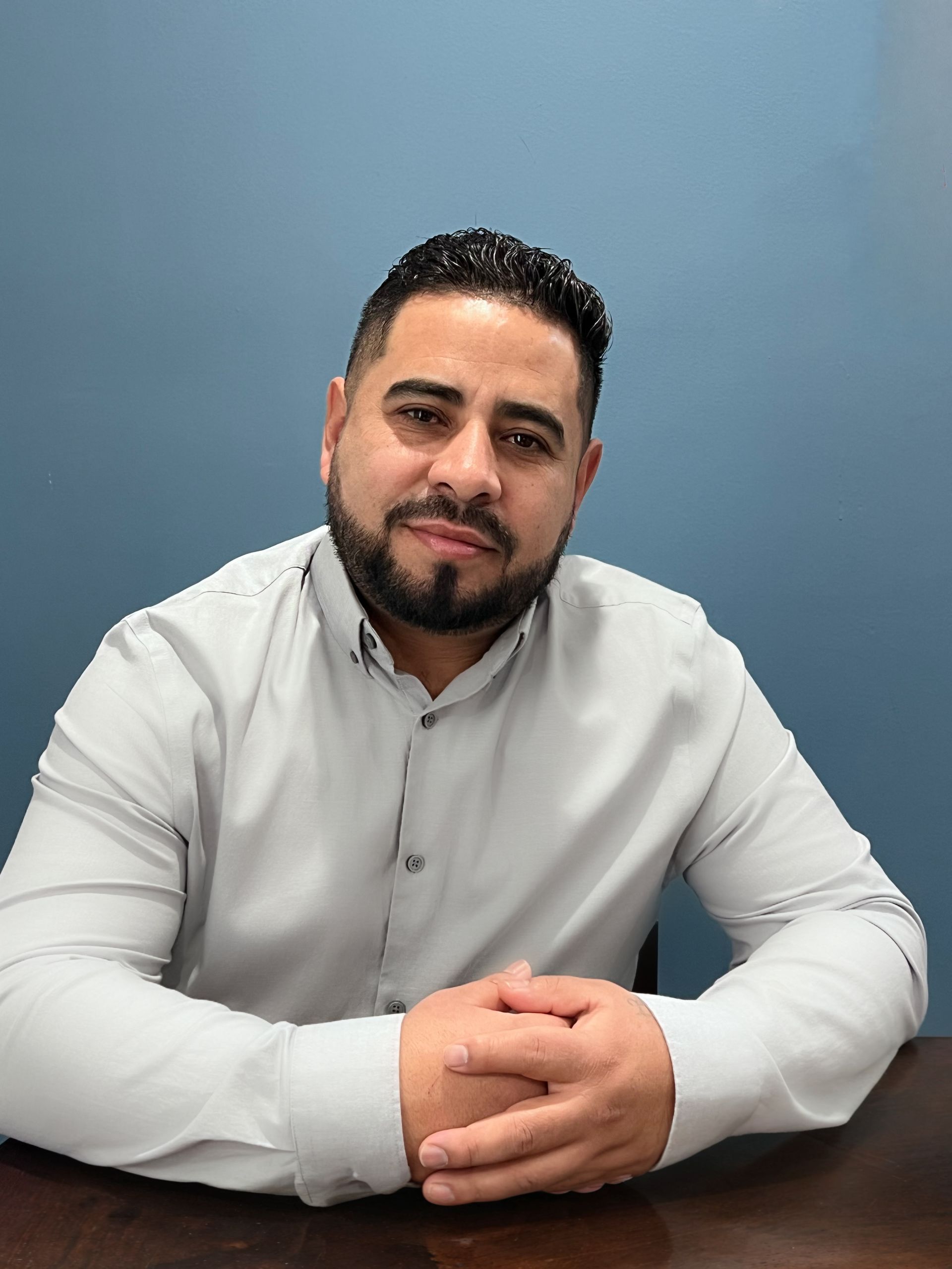Antonio Ortiz with a beard, wearing a white collared shirt, sits at a desk against a blue background with hands clasped.