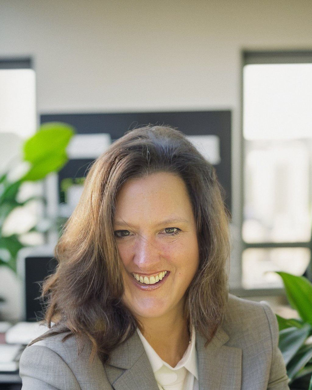 Smiling person in a gray blazer sitting indoors with blurred office plants and windows behind them