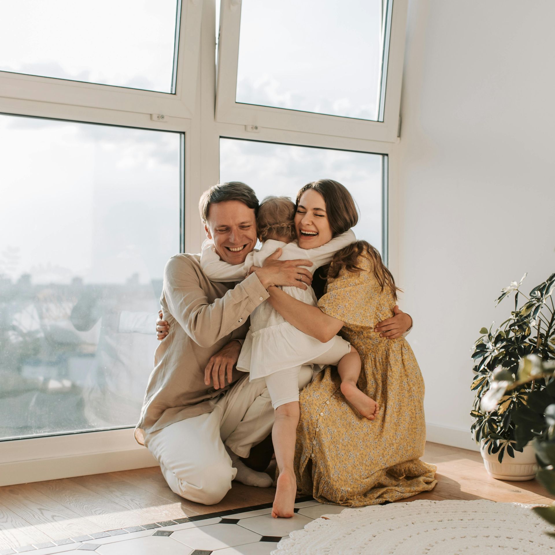 Two people smiling and hugging in a bright room by large windows, with a plant nearby.