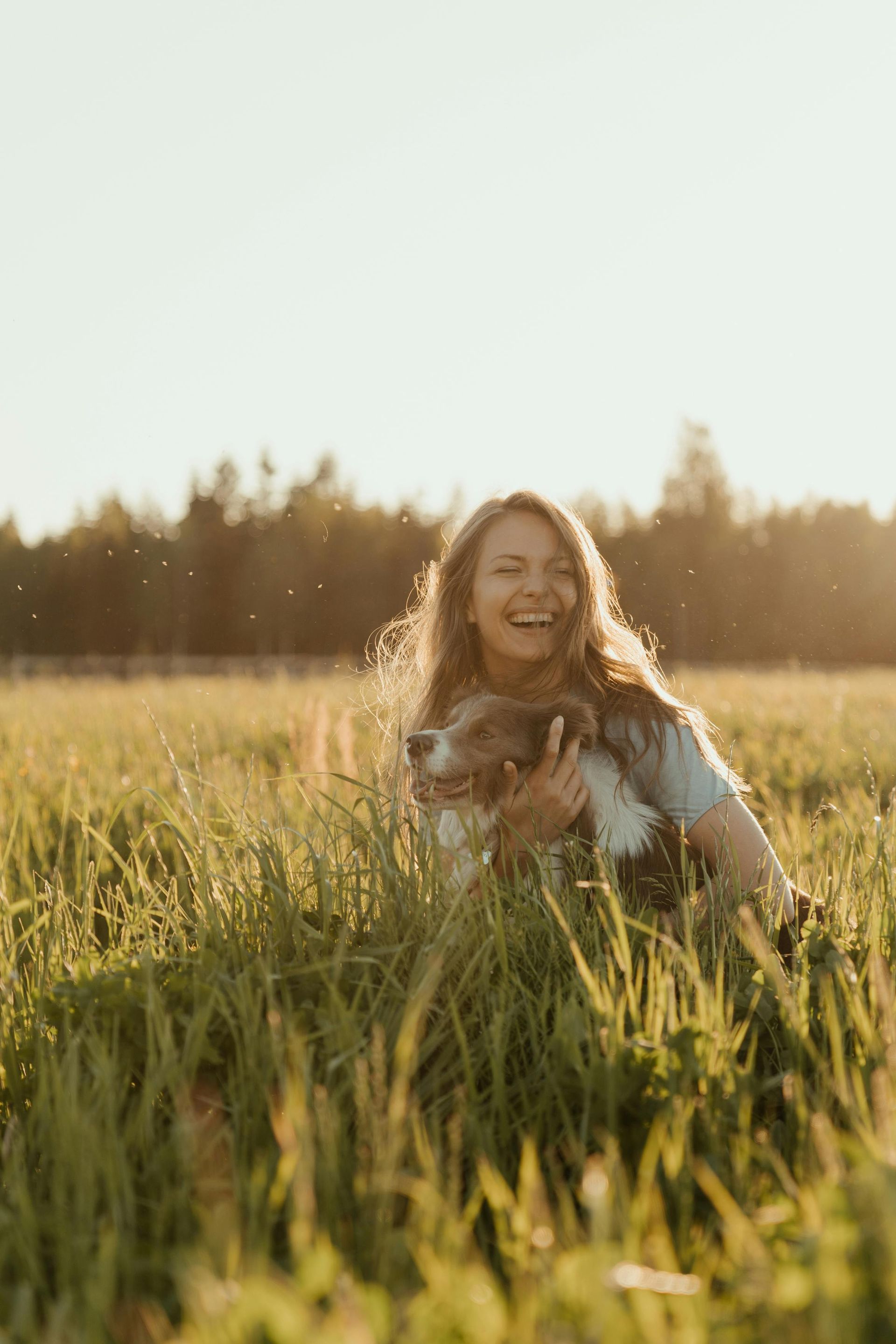 Woman sitting in tall grass at sunset, smiling with a dog in a sunlit field