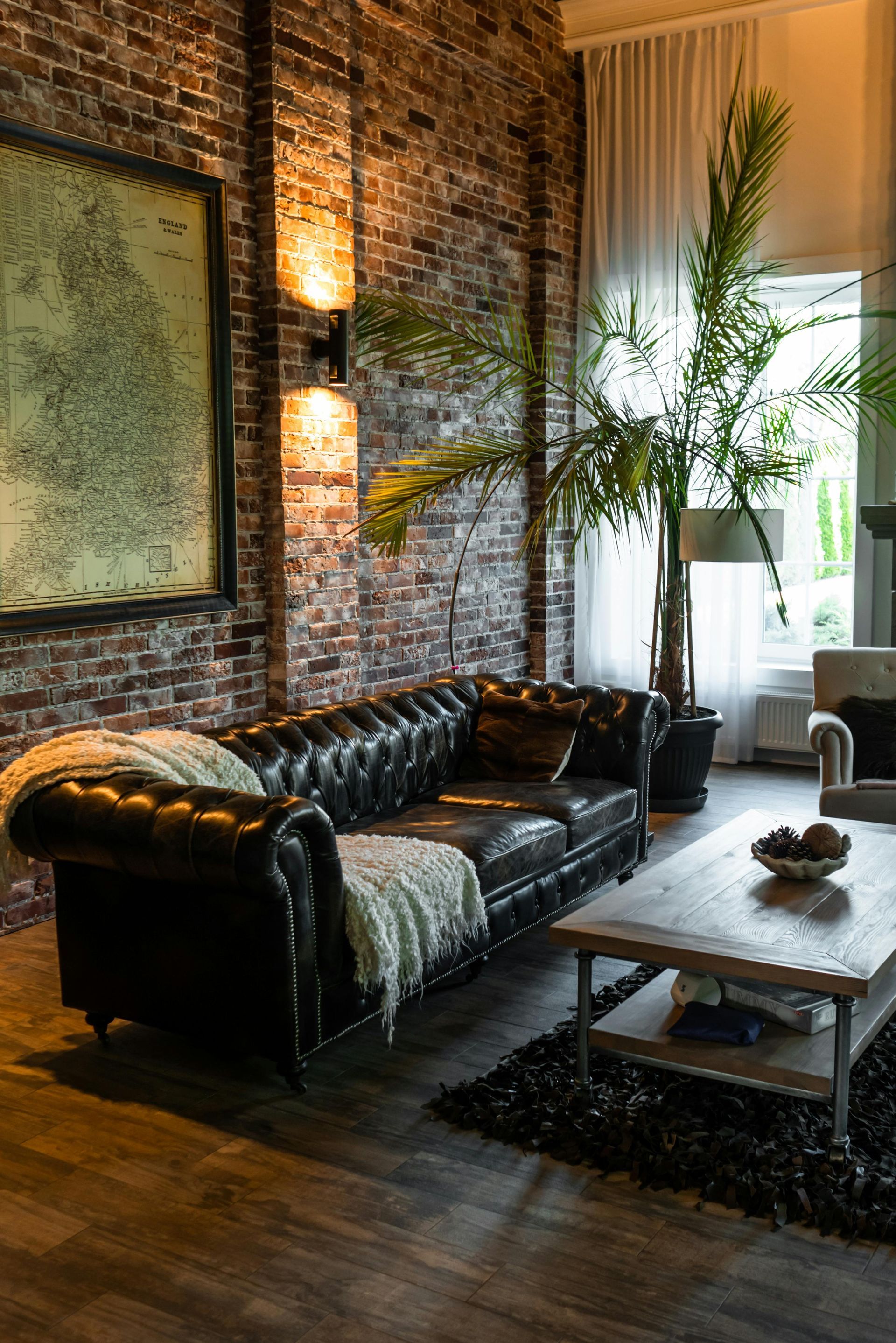 Modern loft living room with black leather sofa, coffee table, brick wall, and tall plants by a window