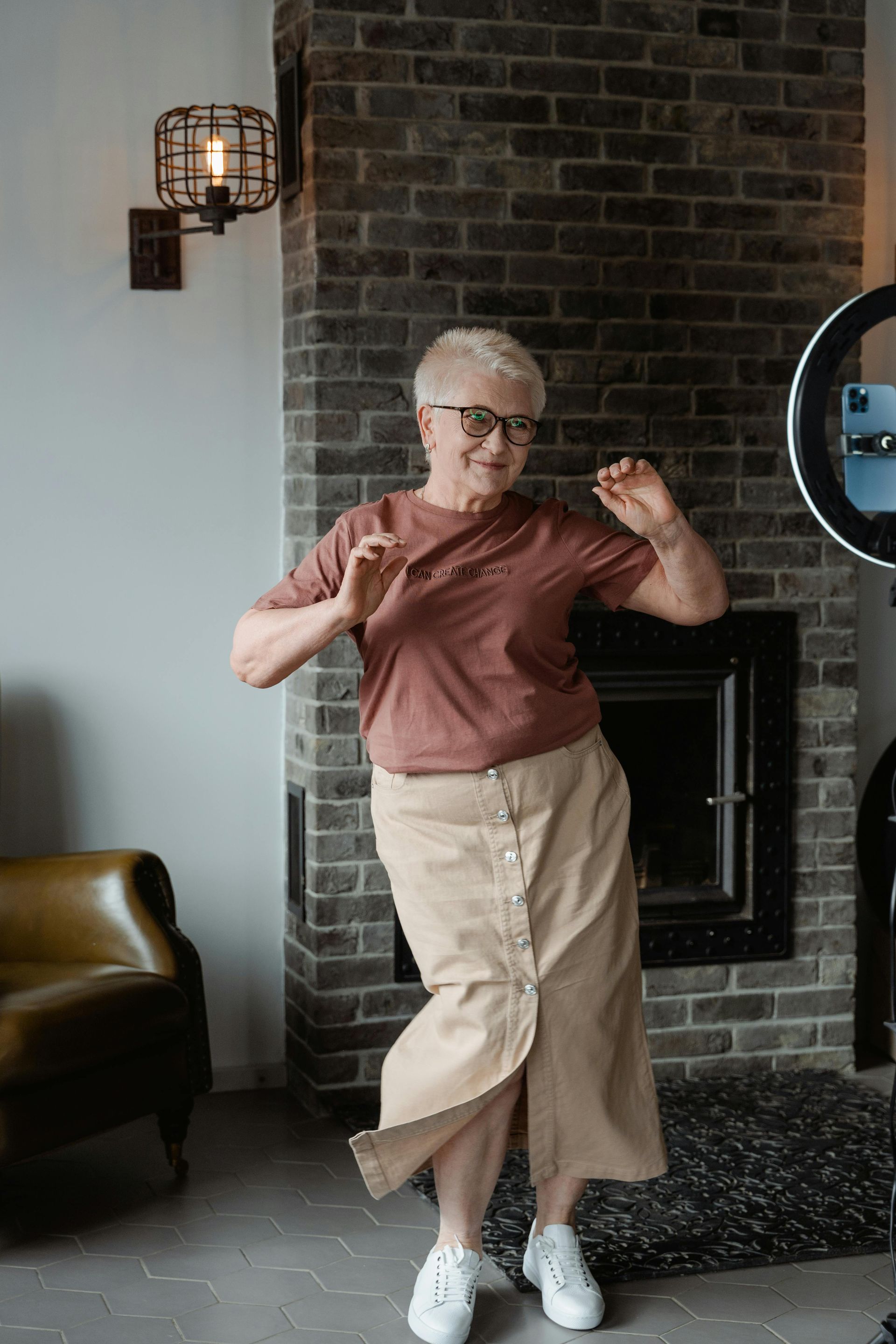 Person posing with hands raised in a living room beside a brick fireplace, wearing a rust shirt and beige pants.