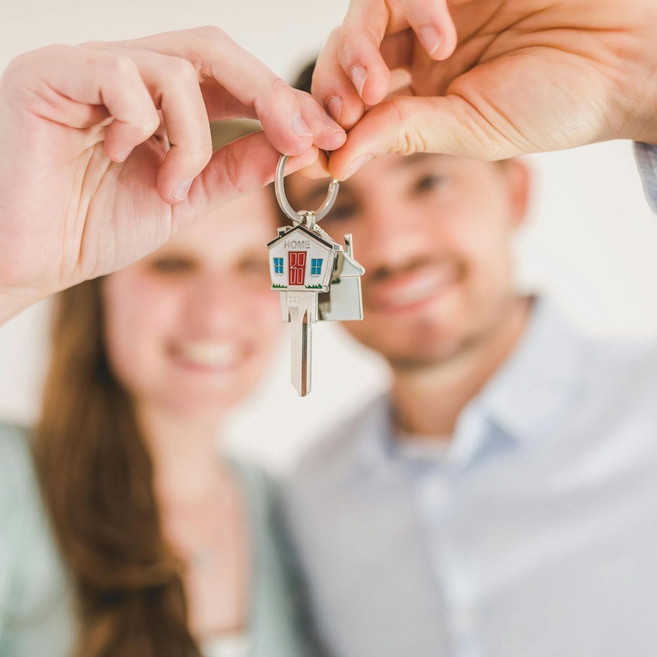 Hands holding house keys with a keychain house, smiling couple blurred in the background