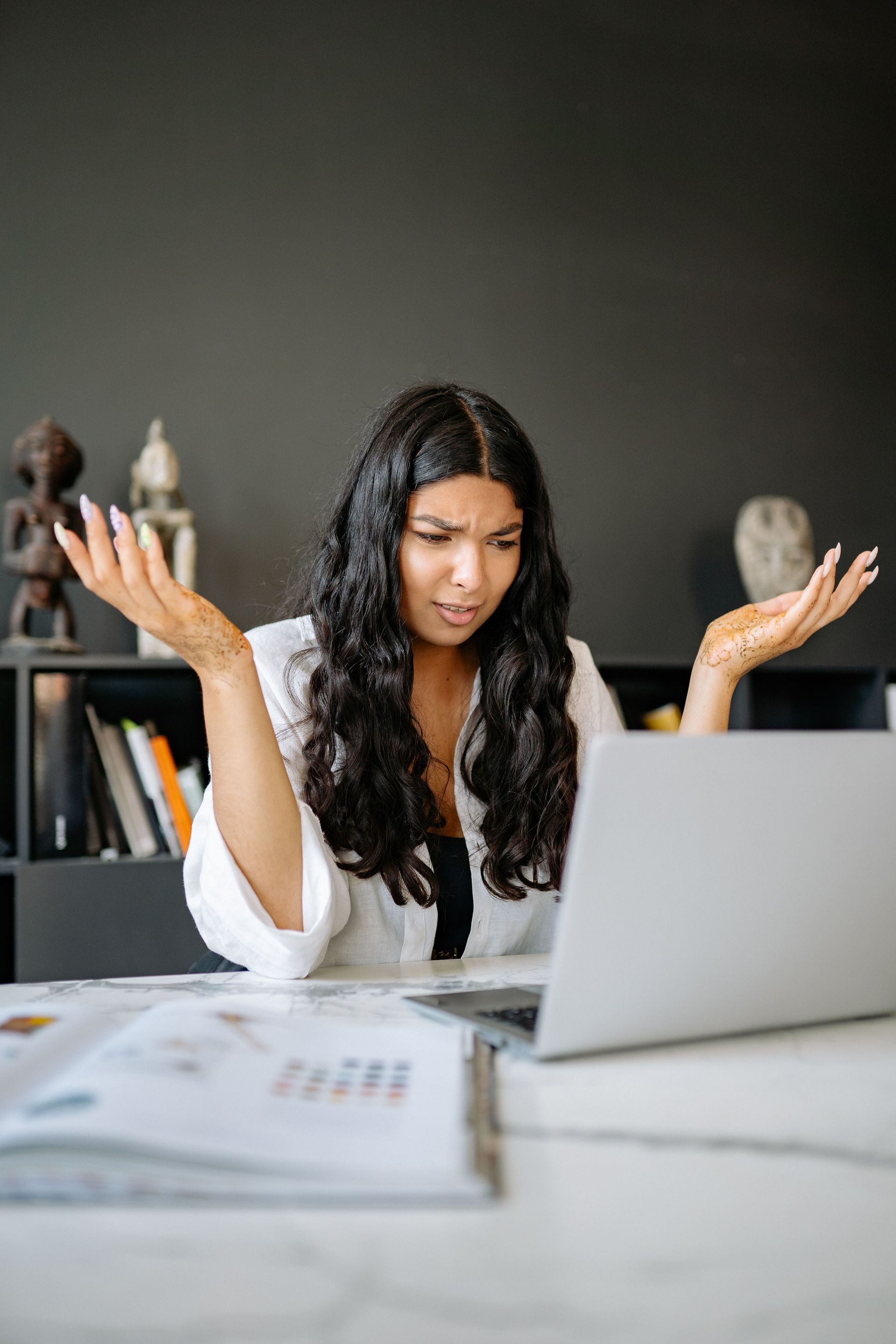 Woman at a laptop with raised hands, looking frustrated at a desk in an office.