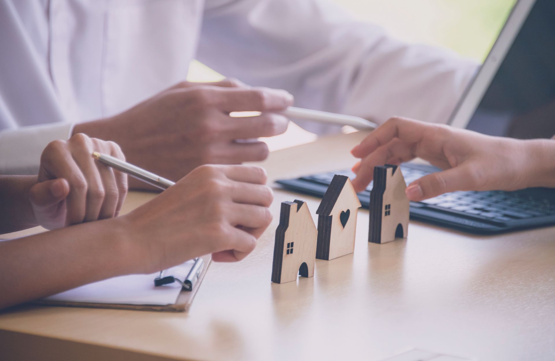 Hands around miniature houses on a desk; people with laptop and clipboard meeting.