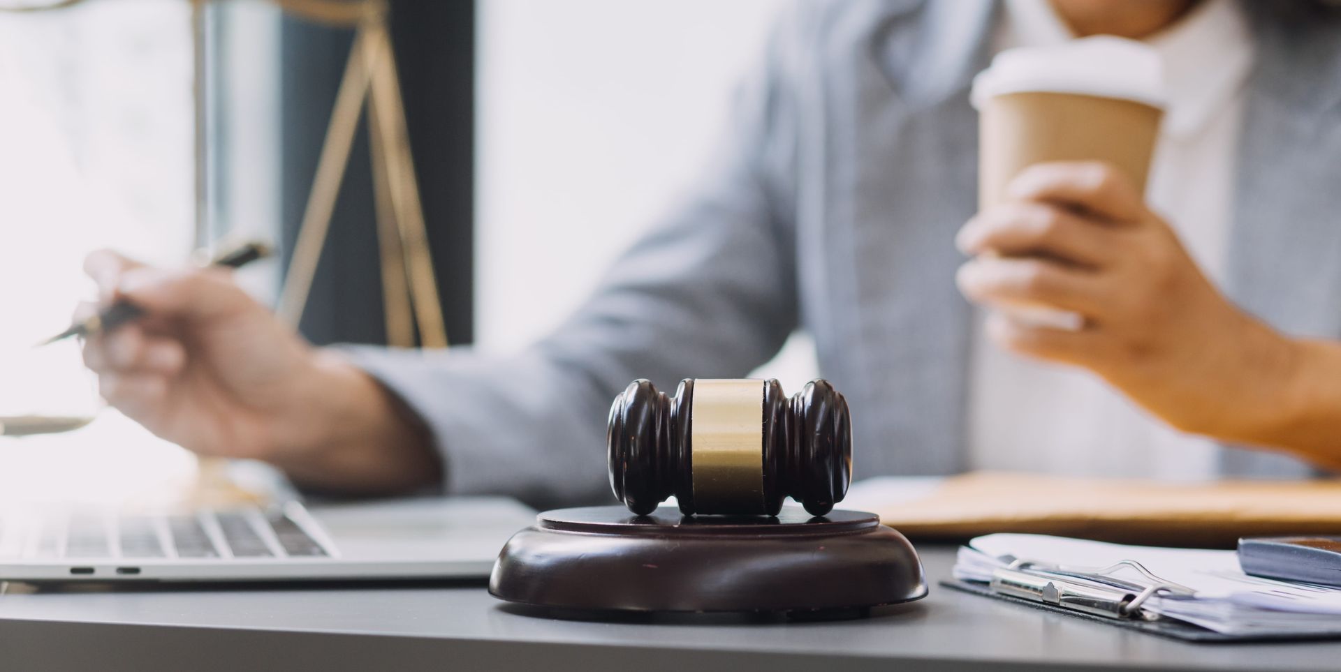 Judge's gavel on desk with laptop and paperwork, while a person holds a coffee in the background