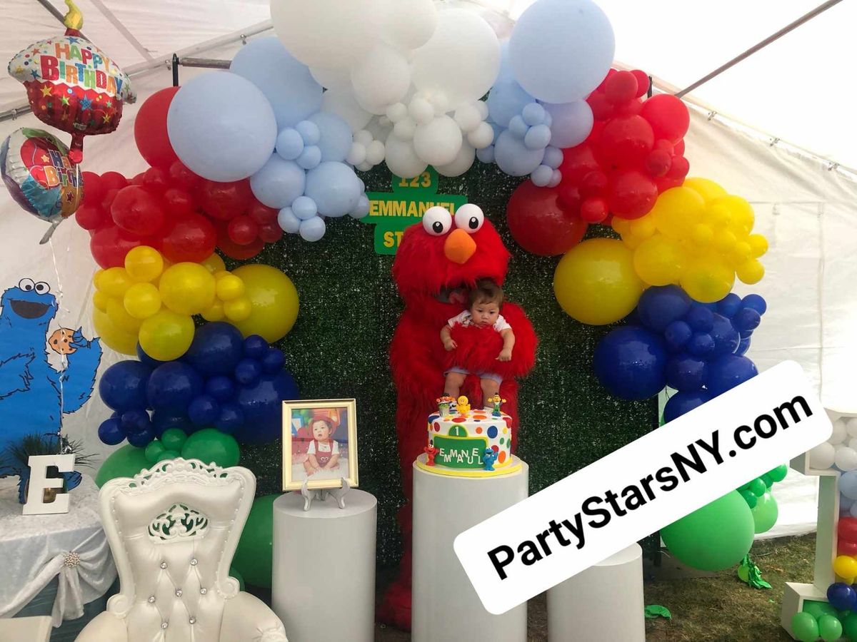 Elmo is standing in front of a wall of balloons.