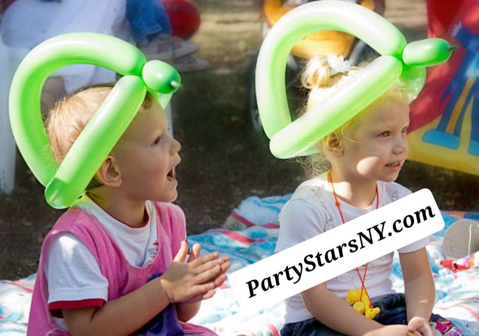 Two little girls wearing green balloon hats are sitting on a blanket ..