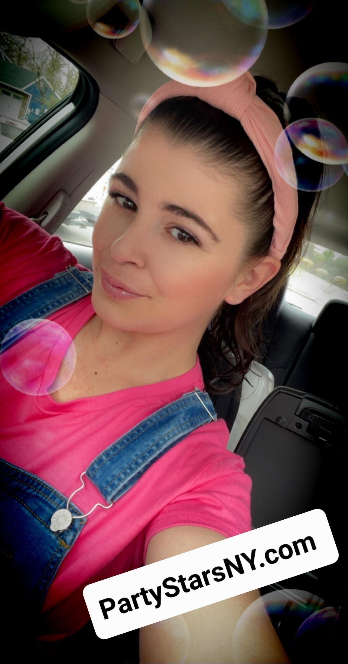 A woman in a pink shirt and overalls is taking a selfie in a car.