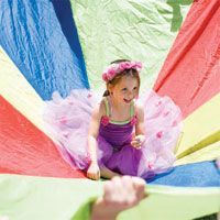 A little girl in a purple dress is sitting under a colorful parachute.
