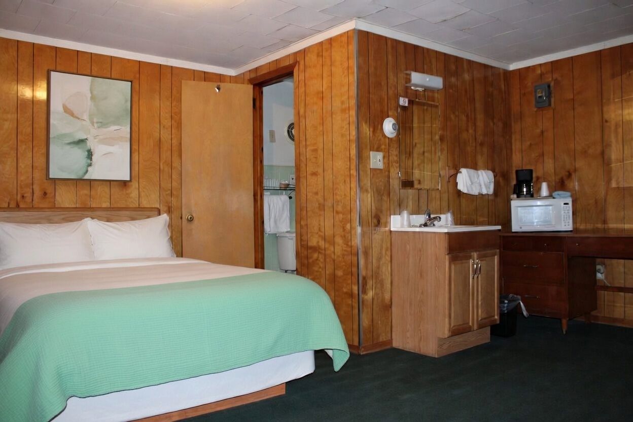 Small wood-paneled bedroom with a bed, mint-green blanket, and a kitchenette on the right.