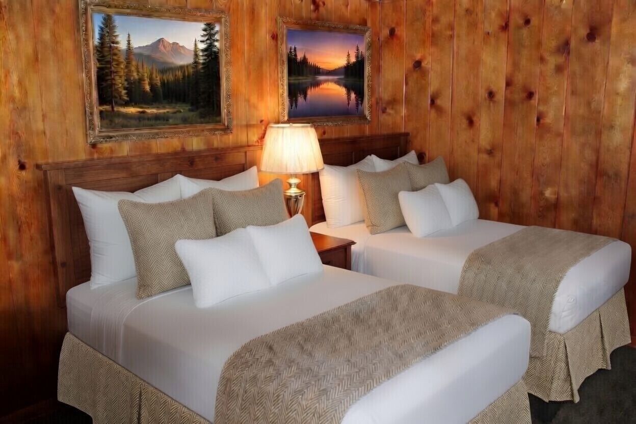 Cozy cabin bedroom with two white beds, wood-paneled walls, a lit lamp, and framed landscape art