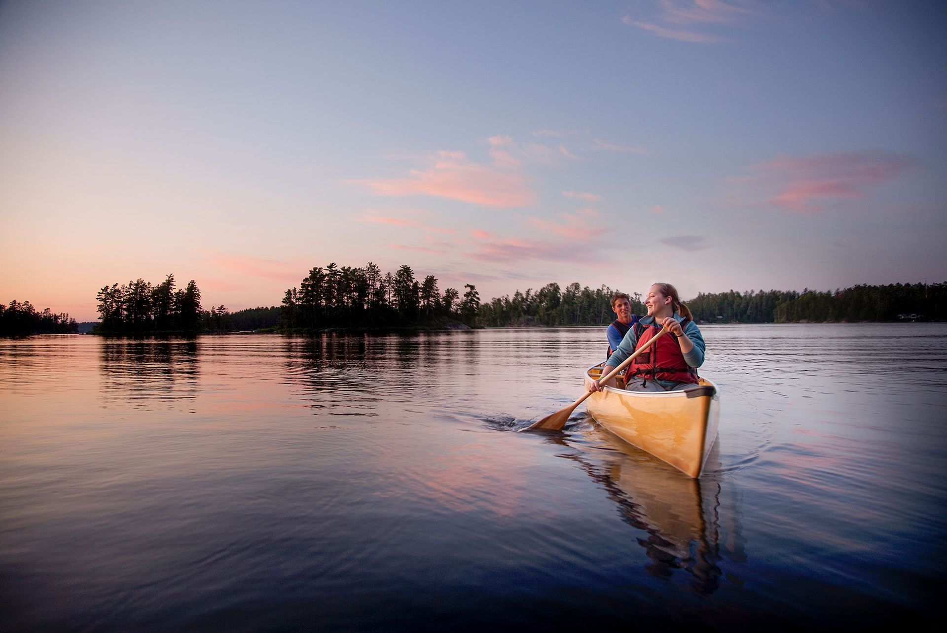 Two people in a canoe on a calm lake at sunset, with trees silhouetted on the horizon.