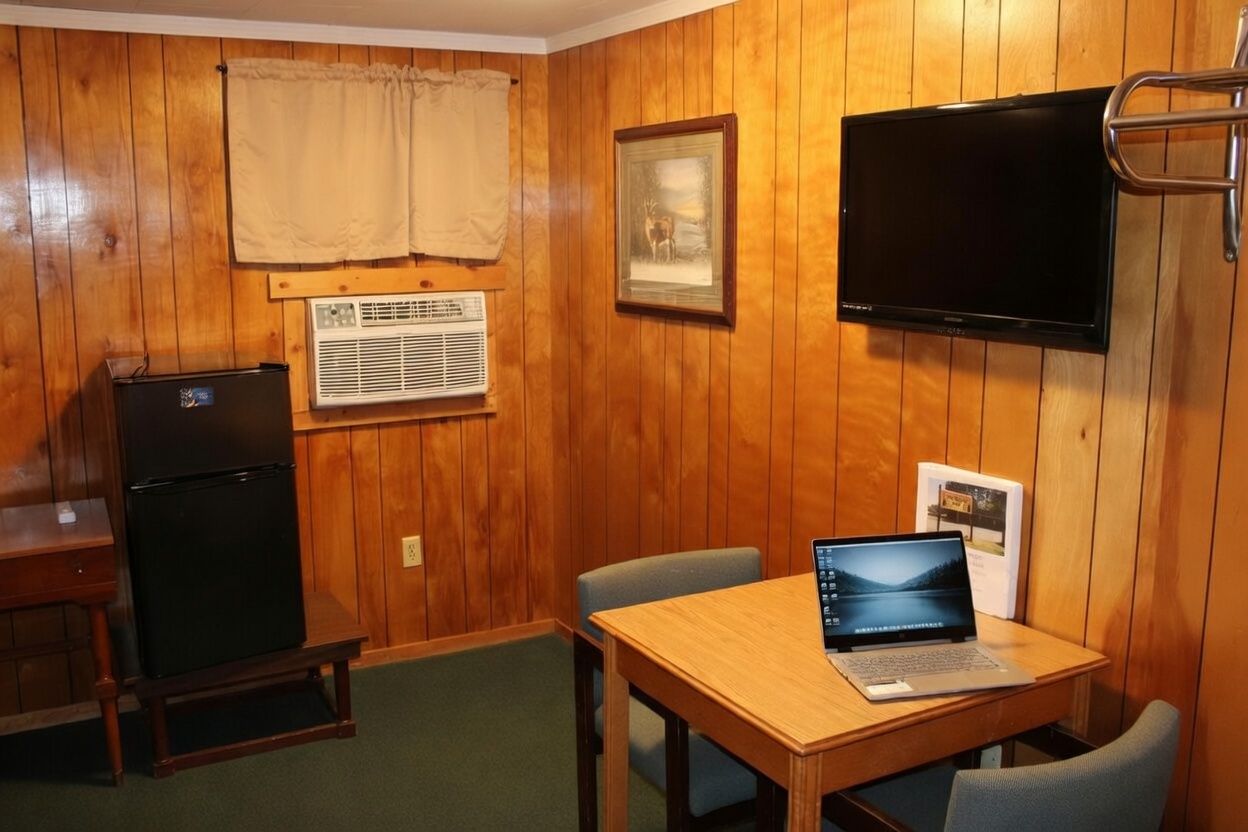 Small wood-paneled room with a TV, laptop on a table, mini fridge, and window air conditioner.
