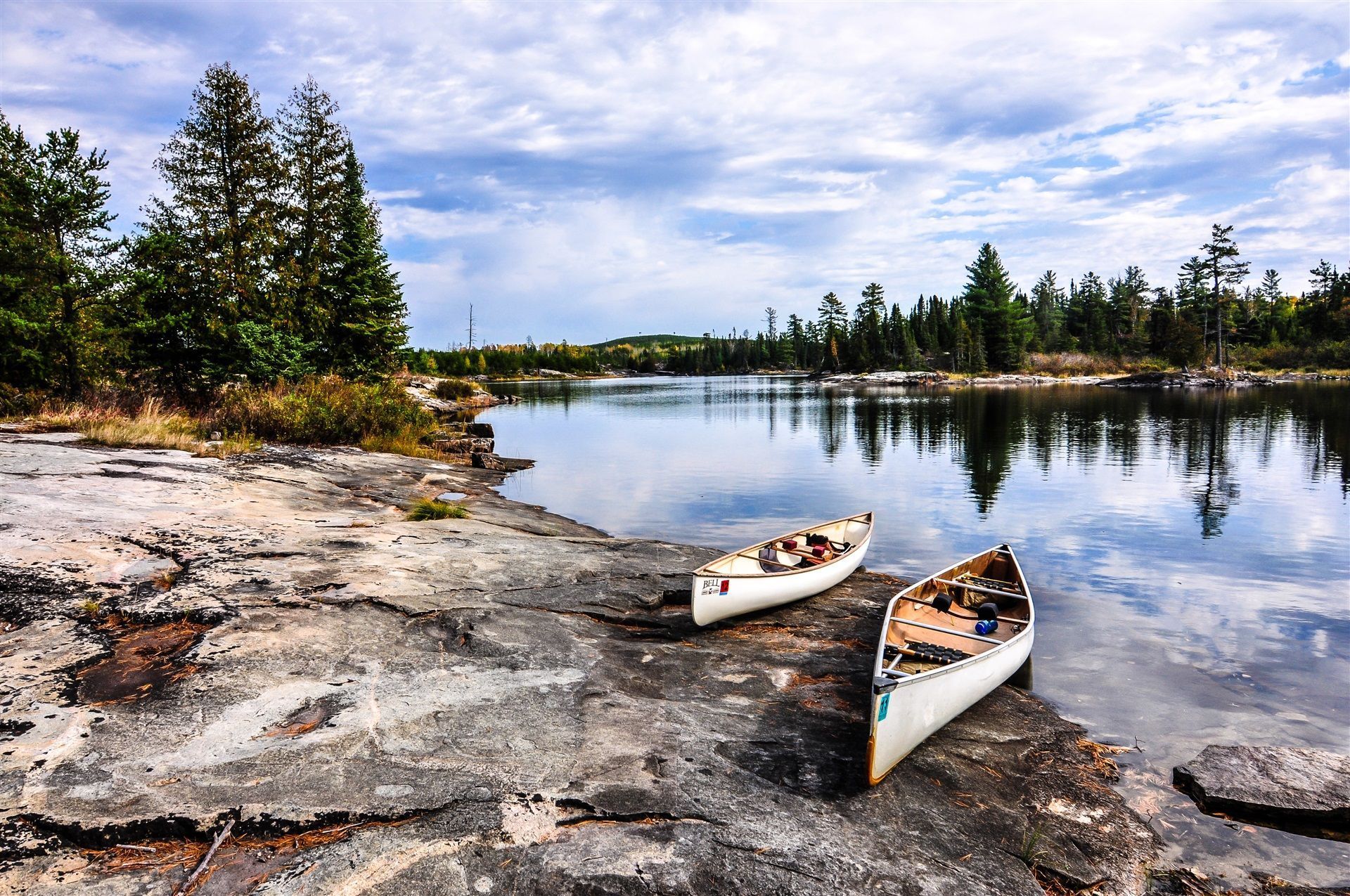 Two canoes on a rocky lakeshore under a cloudy sky, with pine trees and calm water reflecting the shore
