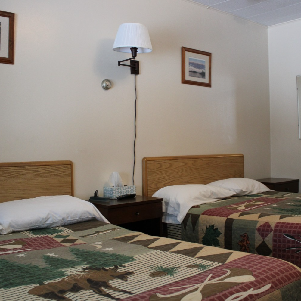 Motel room with two twin beds, patterned bedspreads, and a wall lamp between framed pictures.