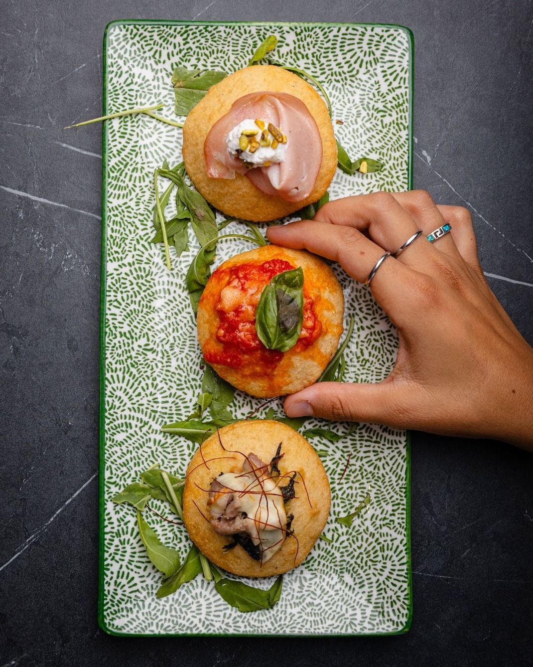 Tre antipasti rotondi di pane piatto conditi con prosciutto, salsa di pomodoro e funghi su un piatto verde. Ne tengo uno in mano.