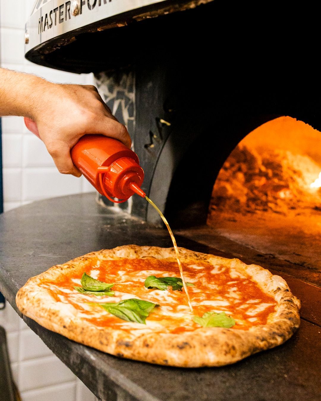 Versare a mano un filo d'olio su una pizza Margherita con foglie di basilico accanto a un forno per pizza.