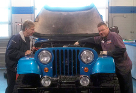 Two men are working on a blue jeep in a garage.