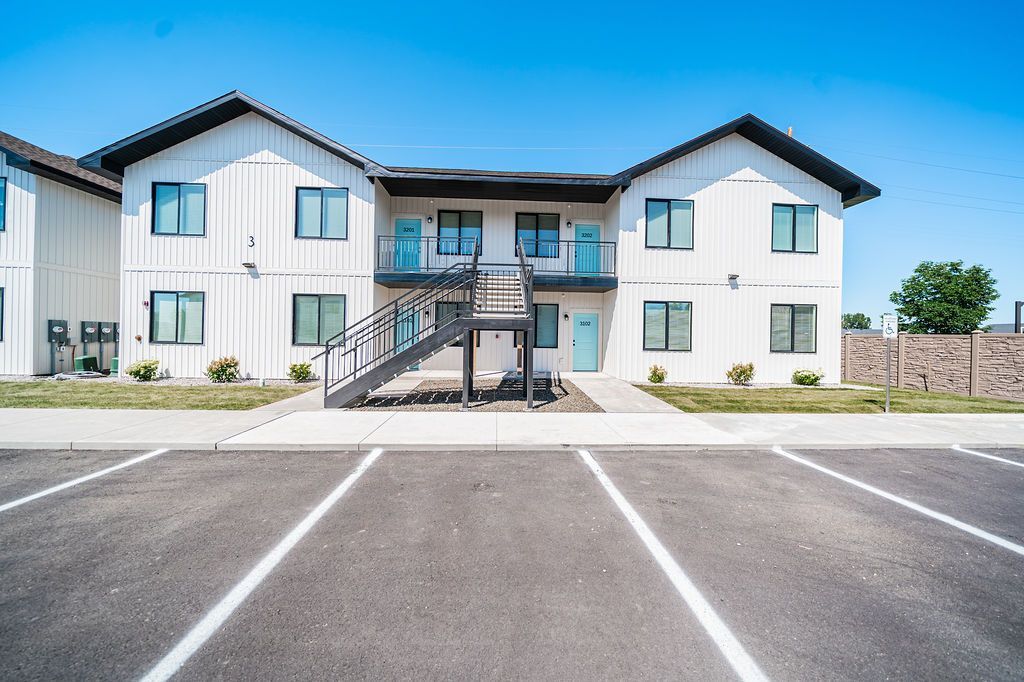 Two-story white building with black trim, teal doors, and a staircase, in front of an empty parking lot.
