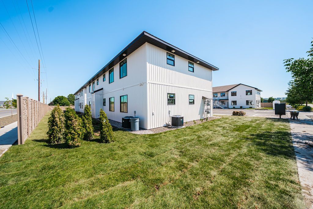 White two-story building with multiple windows, next to a grassy lawn and fence on a sunny day.