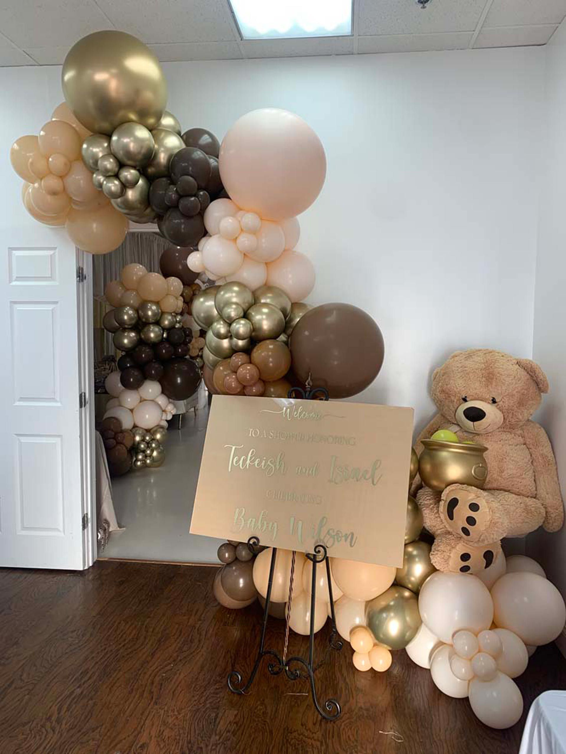 A teddy bear is holding a sign surrounded by balloons in a room