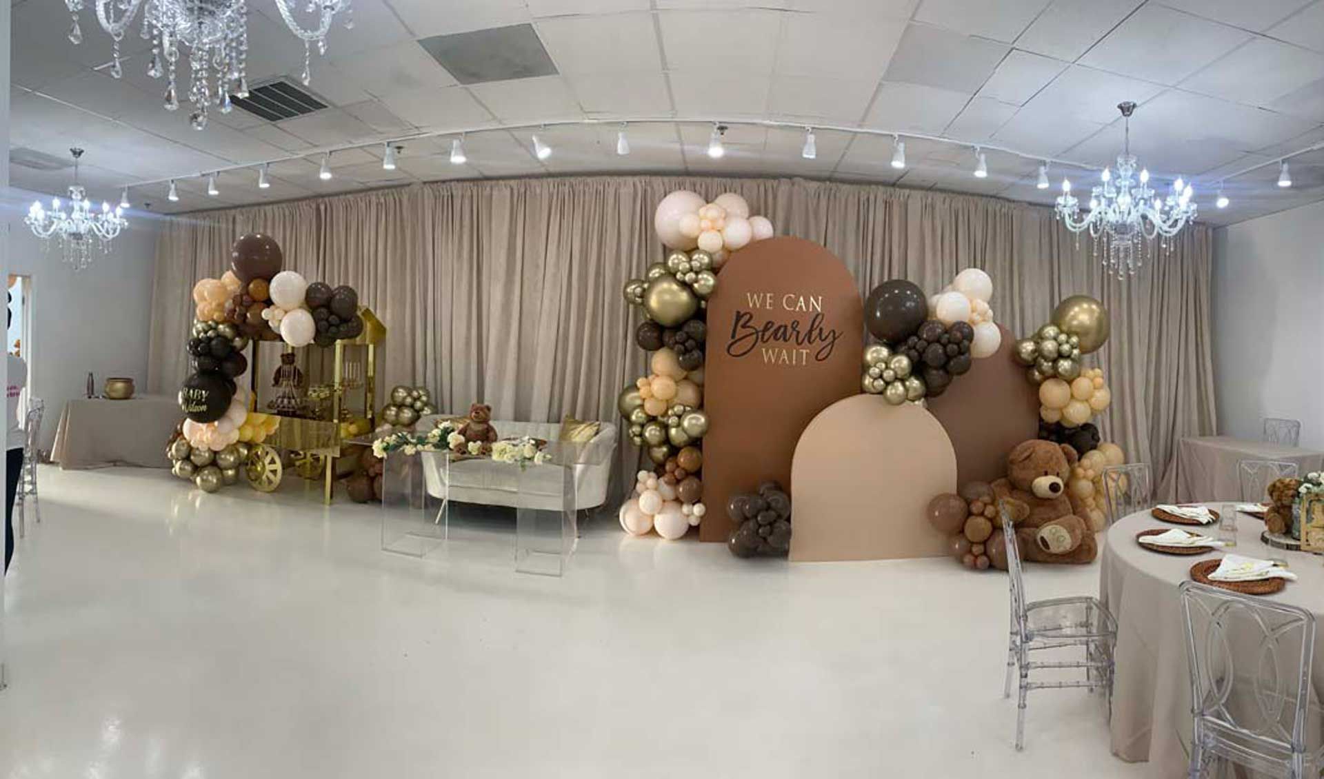 Baby shower with balloon arch, teddy bears, and crib set against a neutral backdrop in a room with chandeliers.
