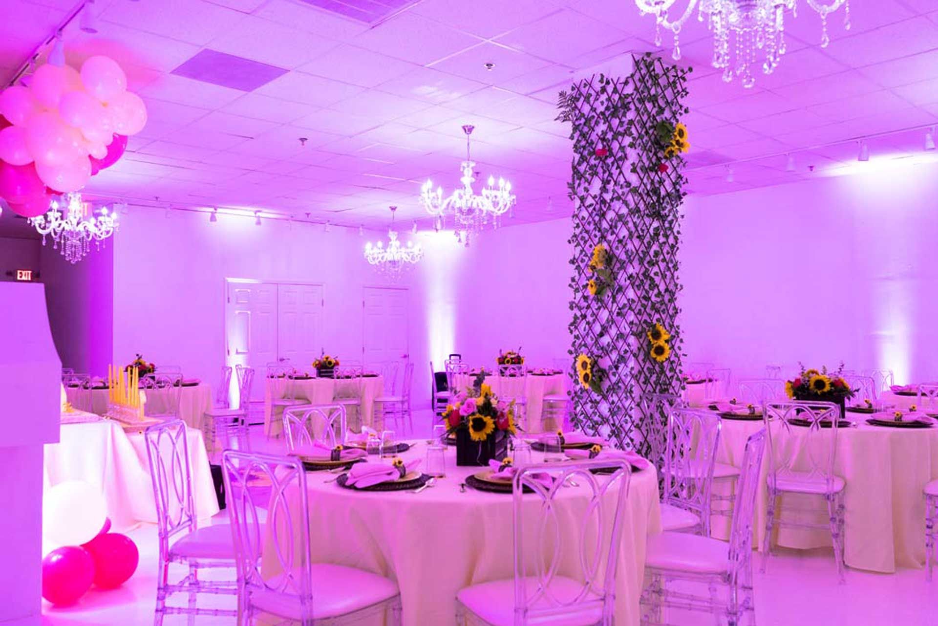Formal event space, round tables with cream linens, clear chairs, purple lighting, floral centerpieces.
