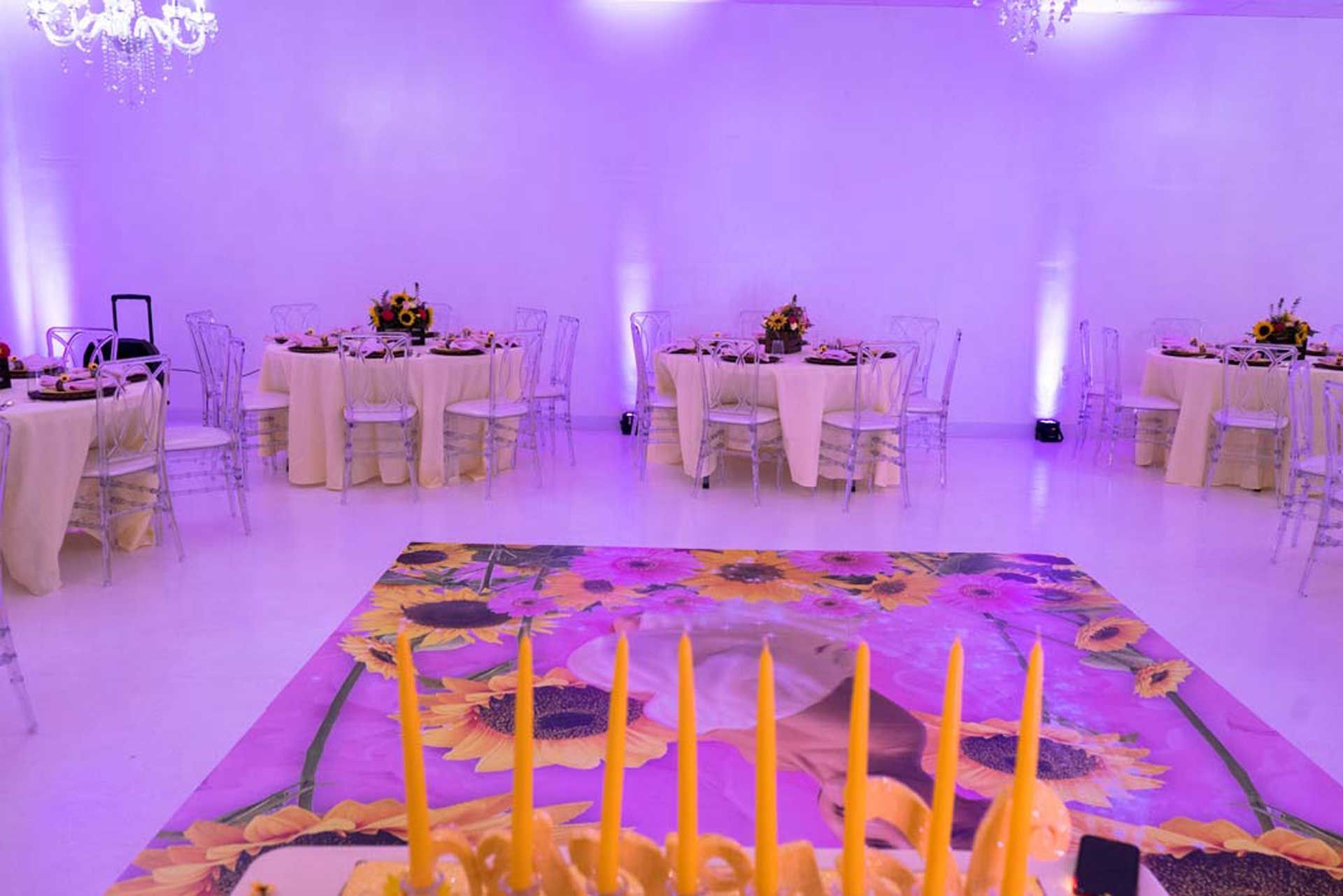 Empty event space with round tables, clear chairs, and a sunflower-themed dance floor, lit with purple light.