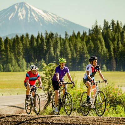 Three people are riding bicycles on a dirt road with a mountain in the background.