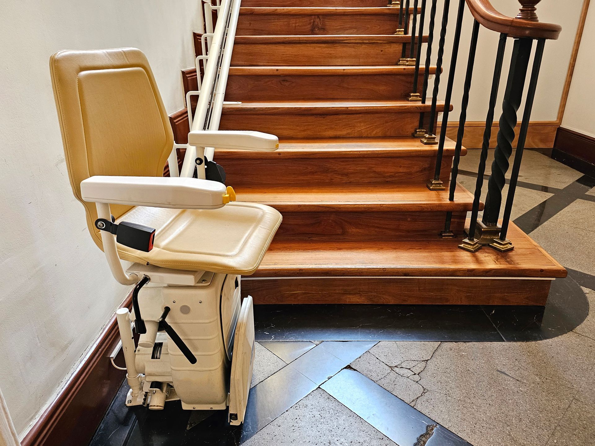 Stairlift on wooden stairs; beige seat, black safety belt, and handrail; indoor setting.