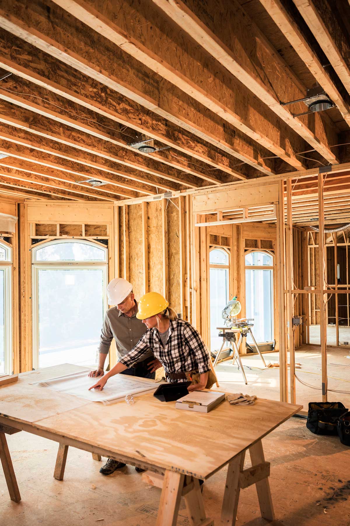Two people in hard hats reviewing blueprints at a construction site. Wooden framing and open windows are visible.