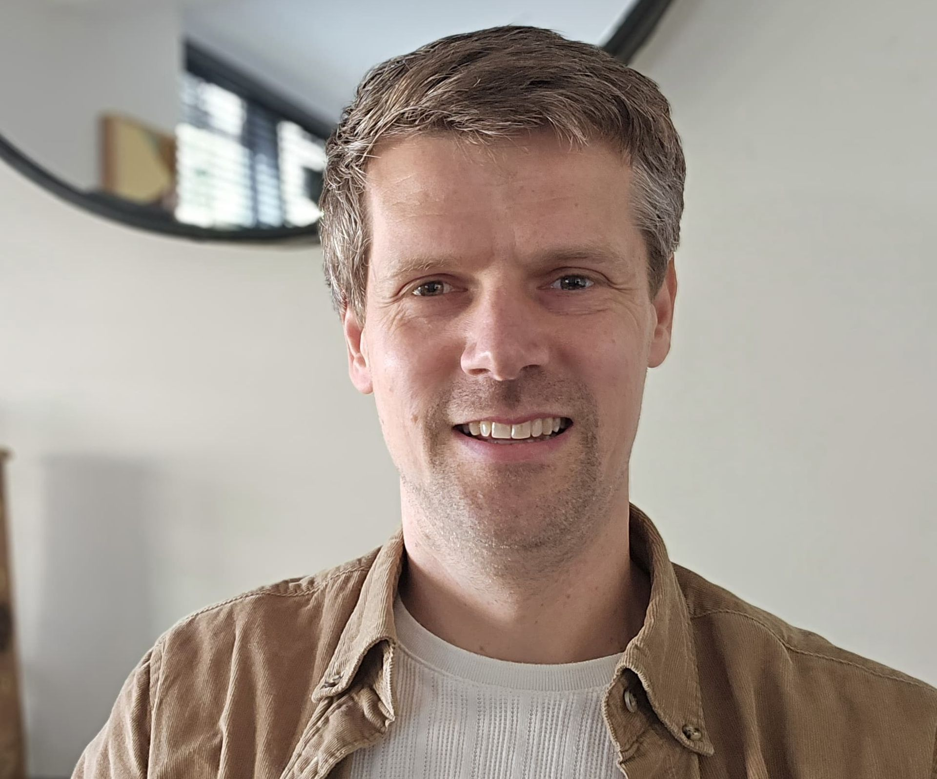 Een persoon met kort bruin haar lacht terwijl hij een beige corduroy overhemd over een wit overhemd draagt, binnen bij een spiegel.