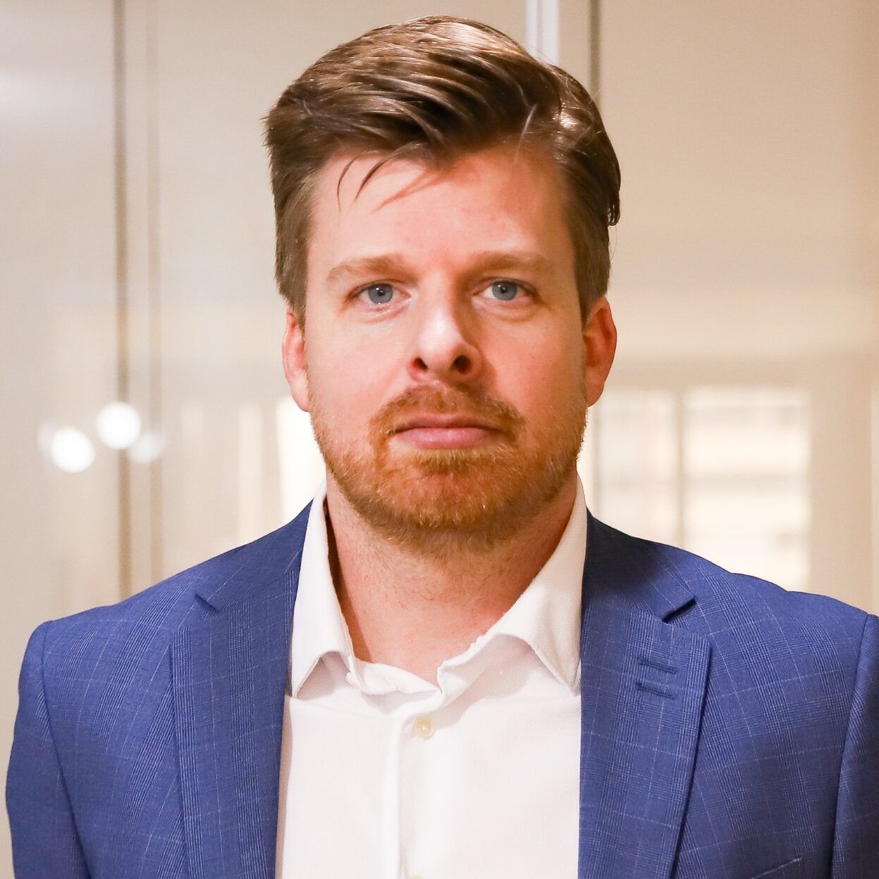 Een persoon met lichtbruin haar en een baard, gekleed in een wit overhemd met kraag en een blauwe blazer, in een professionele omgeving.