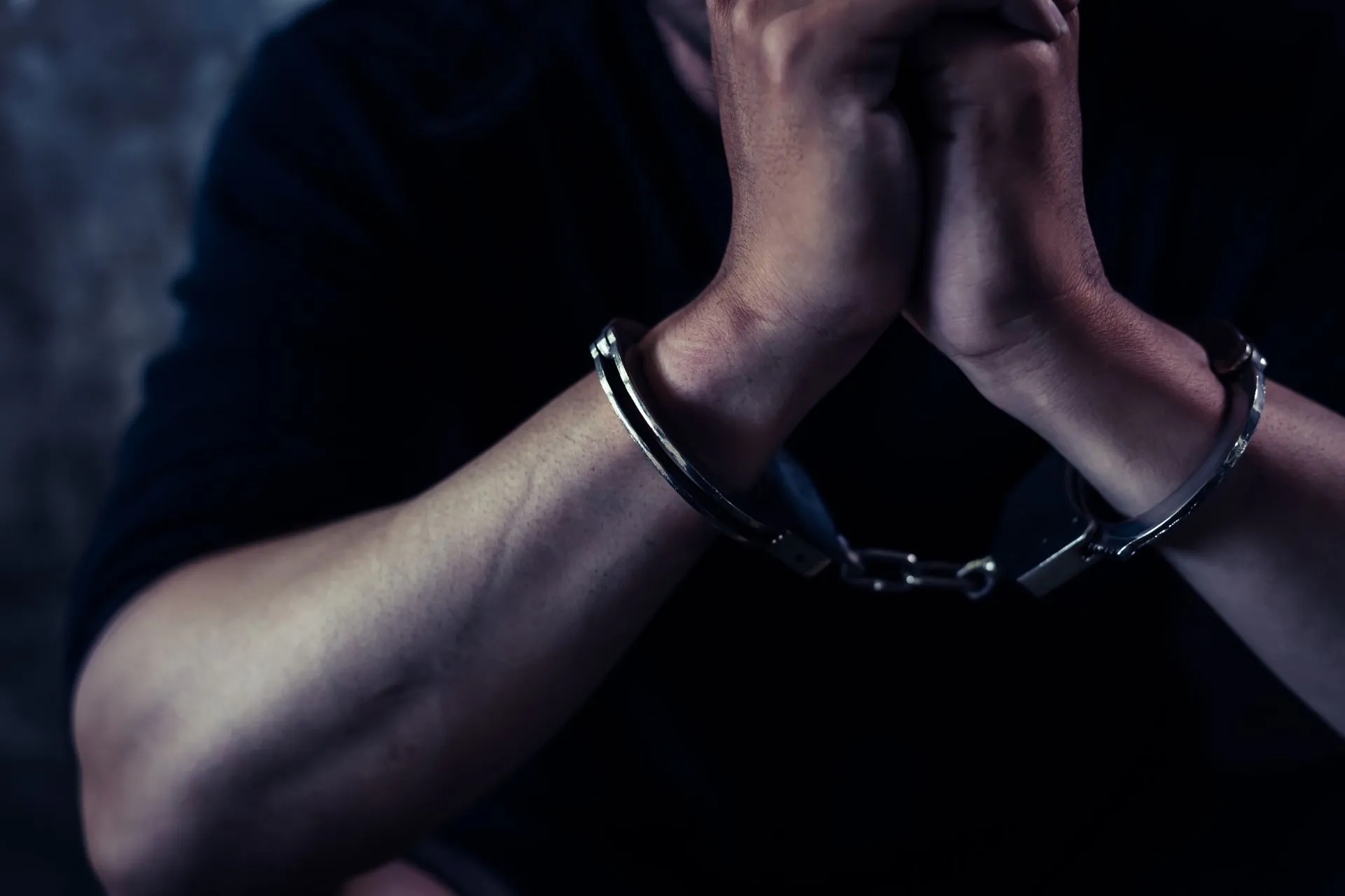 Man's hands clasped in handcuffs, dark clothing, low lighting, indoors.