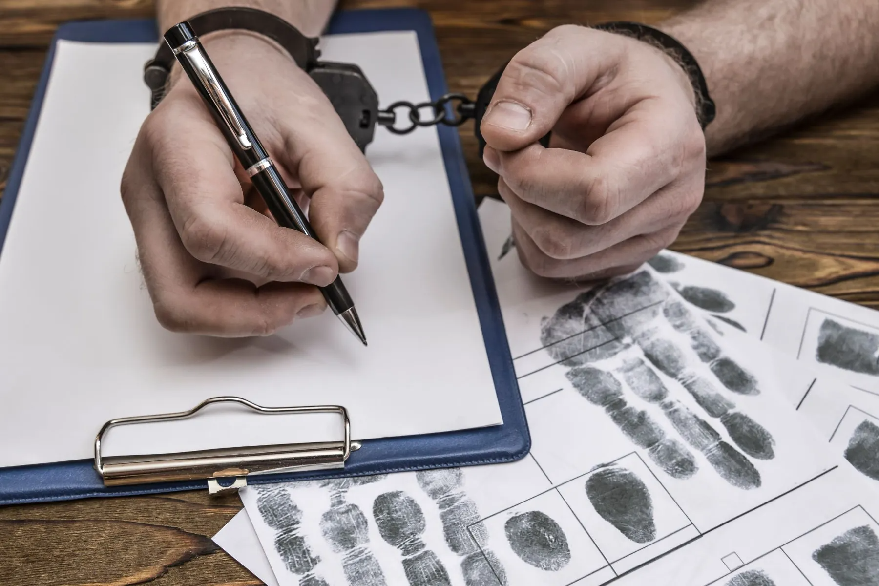 Handcuffed person writing on a clipboard, fingerprints visible nearby.