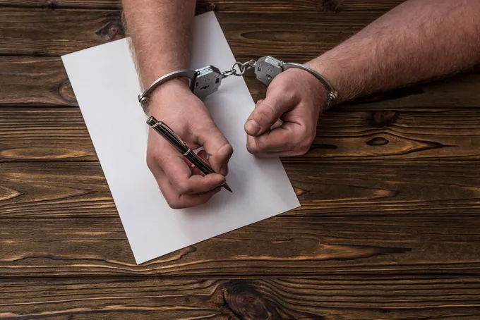 Person's hands in handcuffs writing on a blank paper with a pen, on a wooden surface.