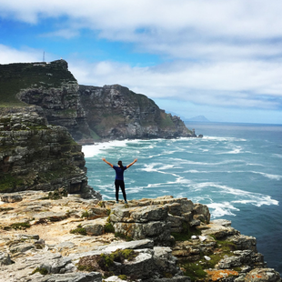 Person standing on a rocky seaside cliff with arms raised, overlooking blue ocean and waves under a cloudy sky