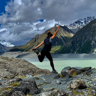 Person posing on rocky shore beside a turquoise mountain lake under cloudy peaks