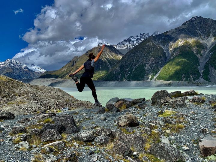 Person jumping on rocky shore beside turquoise lake and snow-capped mountains under a blue sky