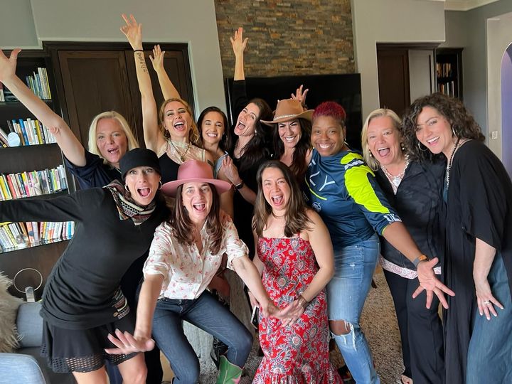 Group of smiling friends posing indoors, arms raised, in a bright living room with stone fireplace and bookshelf