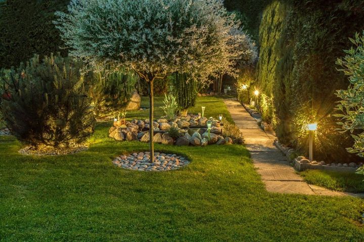 Nighttime garden with lit pathway, lush grass, trees, and a small flower bed with a central pergola
