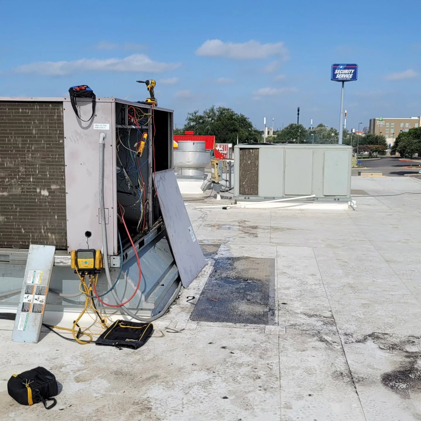 Damaged rooftop HVAC unit with exposed wiring and tools on a flat roof under a blue sky.