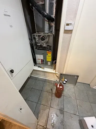 Open furnace closet with exposed HVAC units, tiled floor, and a red fire extinguisher nearby