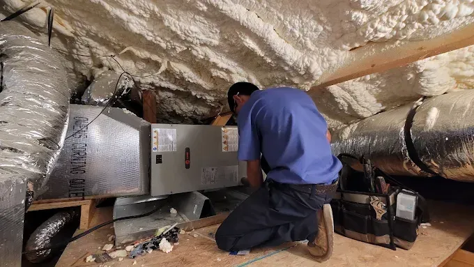 Technician kneeling beside HVAC equipment in an insulated attic, inspecting ductwork and a metal unit